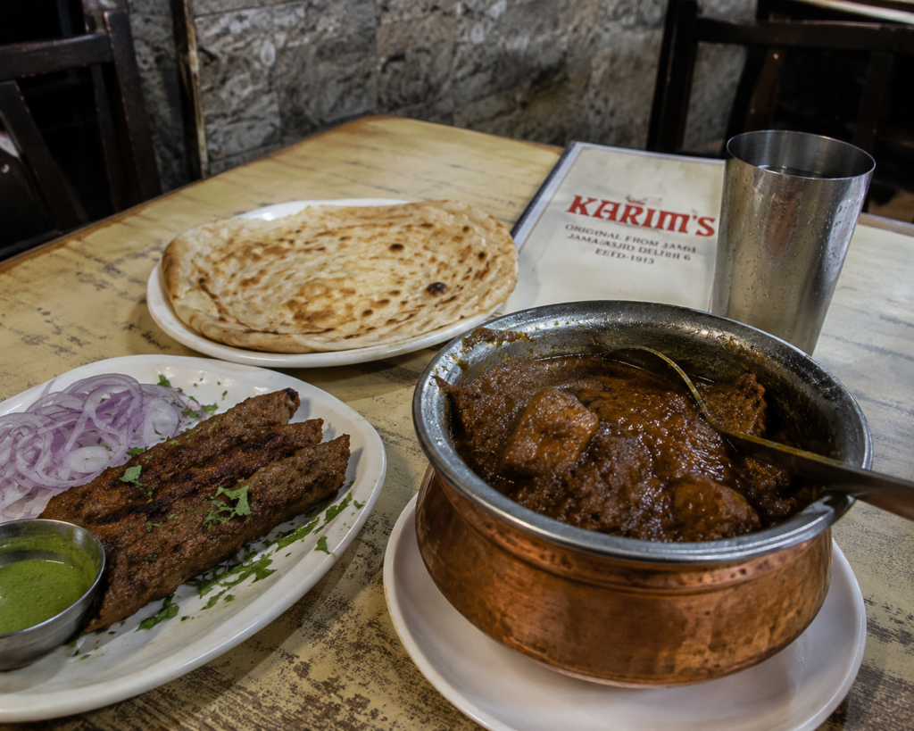 A table at Karim's covered with dishes — a copper pot of dark mutton korma, seekh kebabs- India Food guide 2026 - all the cities popular street food, fine dinning meals, food tours,drinks & desserts.
